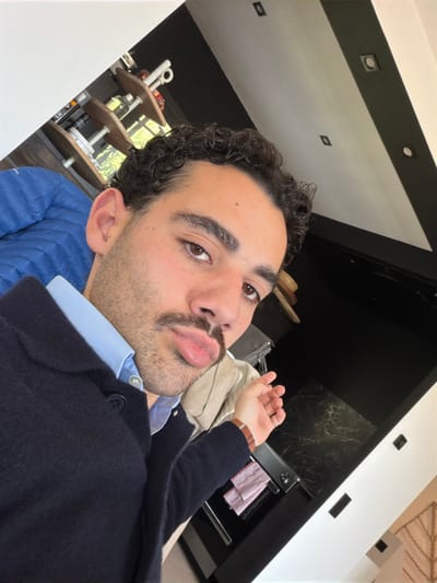 A male individual is depicted in a close-up, angled selfie taken indoors in Mougins, France. The subject has dark, curly hair, brown eyes, and a prominent mustache, and is making a puckered lip expression while looking directly at the camera. He wears a light blue collared shirt under a dark navy blue button-up cardigan. A multi-strand brown leather bracelet is on his left wrist.

The background on the upper left reveals a blue puffer garment and a multi-tiered wooden shelf unit containing several bottles and other objects, with green foliage visible through an opening or window. The background on the right features a contemporary interior with a black wall section, a dark, possibly marbled, countertop, and a modern kitchen appliance with a metallic handle. A red and white striped cloth hangs on a rail. The ceiling is white with dark, linear accents and square recessed lighting fixtures.