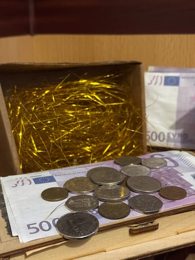 The image presents an overhead view into an open container, featuring a collection of money and golden decorative material. On the right side, a light-colored wooden box is partially visible, showing a simple wooden latch mechanism.
Within this container, a stack of 500 Euro banknotes is prominently displayed, with the "500 EURO" denomination clearly visible. Resting on and around these banknotes is an assortment of various coins. The coins display diverse designs, indicating a mix of international currencies. Visible examples include a British coin featuring the profile of Queen Elizabeth II, an octagonal coin with an indistinct design, and another coin with what appears to be Arabic script, possibly an Omani Rial. Several other coins of varying sizes and metallic finishes, likely Euro coins, are also present.
To the left, a separate section of the container, appearing to be made of brown cardboard, is filled with a dense, shimmering mass of golden tinsel or shredded metallic paper. This material creates a striking contrast with the currency and the wooden elements. The lighting in the image suggests an indoor setting, possibly under artificial light, which accentuates the reflective qualities of the coins and tinsel. No people are visible in the scene. The overall impression is that of a personal collection of money, potentially stored in a decorative or significant manner, such as a treasure chest or savings box.