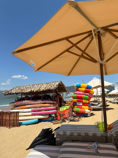 The image depicts a beach scene in Quy Nhon Ward, Vietnam. In the foreground, a large, beige beach umbrella shades the sand. Beneath the umbrella are two lounge chairs with striped cushions, one of which has a water bottle and rope on it. To the left, a stack of colorful stand-up paddleboards leans against a thatched-roof shelter. Behind the paddleboards, several life vests are visible in a wire cage. A pile of inflatable rings, stacked high and brightly colored, stands to the right of the paddleboards. In the background, the ocean stretches out, with gentle waves lapping the shore. Distant buildings and mountains form the backdrop under a clear blue sky with a few wispy clouds. The overall scene suggests a recreational area on a sunny day.