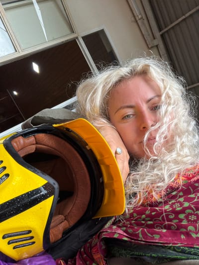 In Hướng Phùng Commune, Vietnam, a close-up shot captures a woman with blonde, curly hair resting her head on a yellow and black helmet. She is wearing a red and green patterned scarf. The background appears to be an open structure with a corrugated metal roof and some windows.