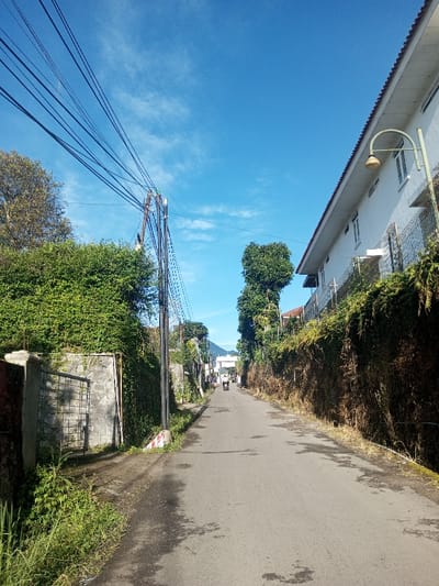 The image displays a narrow, paved street in Khouathineung, Laos, under a clear blue sky with scattered white clouds, indicating full daylight. On the left, a prominent utility pole supports multiple overhead electrical wires that traverse the frame. Below the wires and pole, a stone retaining wall, partially covered by green vines, features a metal gate. Further left, dense green foliage and trees line the street. The immediate left foreground consists of green grass and weeds. On the right side, a white multi-story building with a red-tiled roof is visible, positioned behind a wire fence and a heavily vegetated embankment. A light-colored streetlamp extends from the right side, either from the building or an adjacent pole. The asphalt road surface appears dark, with lighter patches and stains, extending into the distance. In the far background, several indistinct figures, some operating motorcycles, are observed moving away from the viewer down the street.