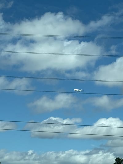 A white airplane is visible in the sky, flying horizontally between power lines. The sky is blue with scattered white clouds. The airplane is small in the frame, suggesting it is at a distance. Power lines run horizontally across the image, obscuring parts of the sky and clouds. The context provided suggests this scene is in Chester, United Kingdom.