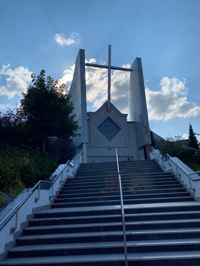 The image shows a wide, grand staircase leading up to a modern church building, likely located in the city of Przeźmierowo, Poland. The church features a prominent cross at its apex, silhouetted against a bright, partly cloudy sky. The architecture is characterized by angular, grey concrete structures. To the right of the entrance, a statue of a religious figure is visible. The staircase itself is made of stone with metal railings on either side. Lush greenery, including trees and bushes, borders the left side of the staircase, adding a touch of nature to the scene. The sun is shining through the clouds, casting rays of light that highlight the cross and the top portion of the church building. The overall impression is one of solemnity and architectural grandeur.