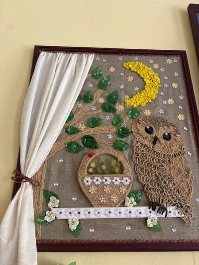 This is a close-up, eye-level, indoor shot of a framed piece of art against a yellow wall. The artwork is a mixed-media collage depicting an owl, a tree with leaves, a crescent moon, a basket with flowers, and a curtain. The owl is made of brown twine, with blue button eyes and a black beak. The tree's trunk and branches are also formed from twine, and its leaves are made of green, translucent plastic. The crescent moon is constructed from yellow ribbon flowers. The basket, woven from twine, contains small fabric flowers. White fabric flowers with purple centers and pink accents adorn a white branch where the owl is perched, along with green plastic leaves. Decorative sequins are scattered throughout the background. The artwork is framed in dark wood and hangs on a textured, pale yellow wall. The specific location within Ветрынскі сельскі Савет, Belarus is not discernible from the image.