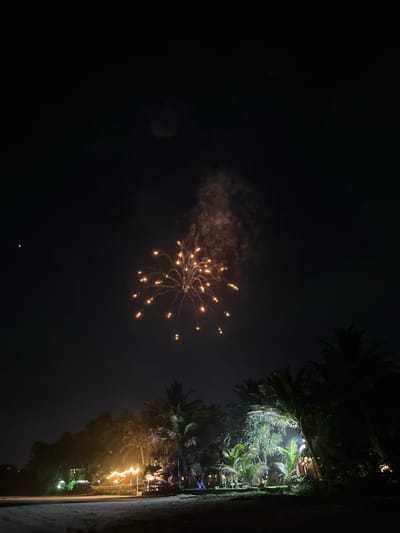 An outdoor nighttime photograph captures a view of a beach and palm trees in Habaraduwa, Sri Lanka. In the upper portion of the frame, a single burst of orange fireworks illuminates the dark sky. To the left of the fireworks, a small, bright, circular object, possibly a planet or star, is visible. The lower half of the image displays a sandy beach in the foreground. Behind the beach, a cluster of silhouetted palm trees and other vegetation is illuminated by strings of warm-toned lights, creating a festive atmosphere. Several small, dimly lit structures are nestled among the trees, suggesting a beachfront resort or restaurant. The overall scene depicts a celebratory night on a tropical beach.
