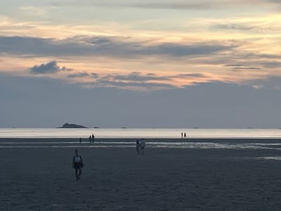 The image shows a beach scene in Baan Sri Thanu, Thailand, at dusk. The sky is filled with layers of clouds, tinged with pale orange hues from the setting sun. The beach is wide and flat, with people walking across it. In the foreground, a woman in athletic wear walks towards the viewer. Further away, a couple walks together, and a small group is gathered near the water's edge. In the distance, a small island is visible on the horizon.