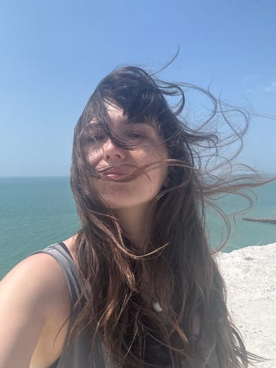 A woman is taking a selfie on a sunny day at the cliffs of Seaford, United Kingdom. Her long, dark hair is blowing in the wind, obscuring parts of her face. She is wearing a grey tank top. The background shows the blue sea and the white chalk cliffs.