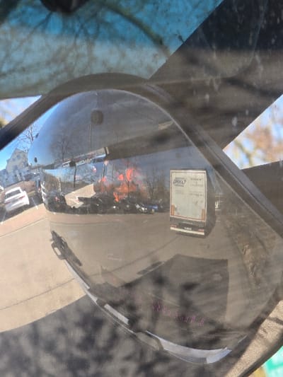 The image depicts a reflection within a convex side-view mirror of a vehicle, capturing an outdoor scene under bright sunlight. The primary reflected environment is an industrial or commercial paved lot, likely asphalt, exhibiting strong shadows indicative of a clear sky.

In the reflected scene, a white box truck or rigid lorry is prominently visible, positioned with its rear facing the viewer. The truck bears a white sticker with "RKL" and an associated logo at the top, and the word "SCHROTT" (German for 'scrap') followed by the number "1328003" below. To the left of the truck, other vehicles are parked, including a white car. Further left, a light-colored building with multiple windows is reflected. In the mid-ground reflection, behind the parked vehicles and to the right of the building, a large pile of what appears to be discarded materials, possibly scrap metal or construction debris, exhibits orange and red hues. Bare trees are reflected in the background, suggesting a non-foliated season.

The mirror's dark gray frame is visible in the foreground, with various streaks, smudges, and minor debris on its surface or the surrounding vehicle window. Faint pink or purple text-like markings are discernable on the lower portion of the mirror's reflective surface.

Based on the "RKL" branding, the text "SCHROTT," and the industrial setting, the scene is consistent with a waste management or recycling facility. This location is identified as Dessauer Str. 129, 06886 Lutherstadt Wittenberg, Germany, specifically a depot or yard associated with RKL Umweltservice GmbH or a related logistics entity.