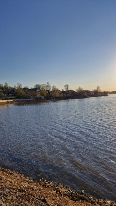 The image depicts a waterfront scene in Otchor, Russia. The sky is clear blue with a hint of light at the top. The body of water extends to the right, showing gentle ripples on the surface. A shoreline with houses and trees runs along the left side of the water. The right edge of the image shows a rocky shoreline. There are no people visible in the image.
