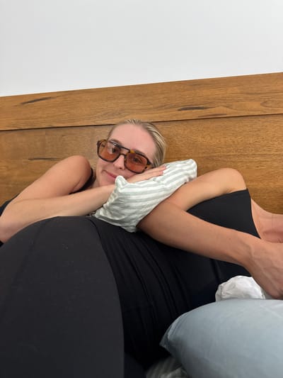 A person wearing orange-tinted glasses lies on a bed with their head propped on a striped pillow. The individual is wearing a black tank top and leggings. A larger, light blue pillow sits behind them. A wood-paneled wall is seen to the left, while a plain white wall is in the lower left corner. The photo is rotated clockwise.