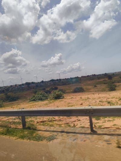 The image depicts a landscape scene in Hoa Thang, Vietnam, characterized by a cloudy sky and a rural environment. In the foreground, a metal guardrail runs horizontally across the frame, partially obscuring the view of the terrain beyond. The ground is a mix of sandy soil and sparse vegetation. In the middle ground and background, a series of wind turbines are visible, suggesting a wind farm in the area. These turbines are spread across a rolling landscape dotted with scattered trees and some buildings, indicating a sparsely populated area with some development. The overall impression is one of a wide-open, somewhat arid landscape under a dynamic, cloud-filled sky. There are no people or specific actions clearly discernible in the image.