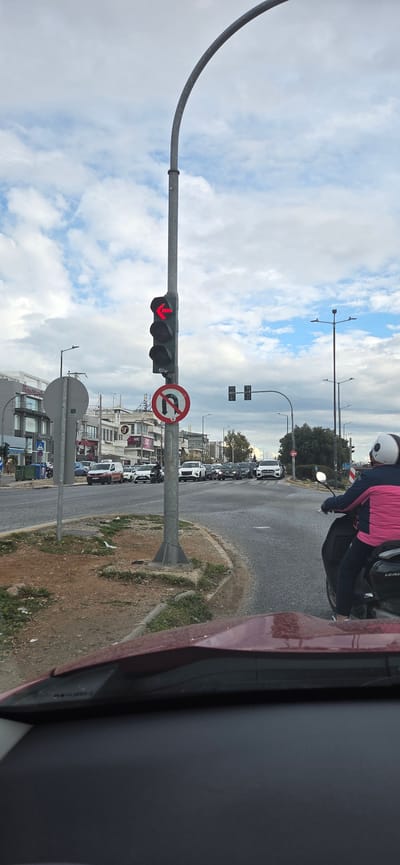 The image presents a street-level view from within a vehicle at an urban intersection in the Municipal Unit of Elliniko, Greece, under a cloudy sky. The immediate foreground shows the top edge of a dark red car's hood and dashboard. A prominent grey pole stands in the left midground, supporting a traffic light displaying an illuminated red left-turn arrow, with a circular 'no U-turn' regulatory sign affixed below it. Directly adjacent to the pole is an unkempt patch of brown earth with sparse green vegetation. To the right, a person wearing a white helmet, a pink and dark blue jacket, and dark trousers is positioned on a black scooter, facing forward in the traffic lane, slightly ahead of the camera's vantage point. The scooter has "LEAD" visible on its lower side. Across the intersection in the background, multiple vehicles, including cars and SUVs of various colors, are stopped in traffic, indicating a queue. Commercial buildings with multiple stories, balconies, and various business signs line the street on the left side of the background. Additional streetlights and some trees are visible further down the road to the right.