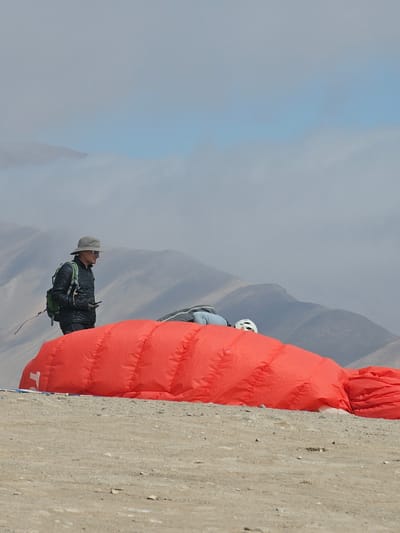 The image depicts two people preparing for a paragliding or hang gliding activity in a mountainous location, presumably in or near Alto Hospicio, Chile, given the city's mention in the query. One individual is wearing a hat and dark clothing and appears to be adjusting gear. A second person, wearing a white helmet, is partially visible behind a large red, possibly inflatable, object. The background shows mountains partially obscured by clouds or fog. A concrete wall or surface is visible on the right side of the image.