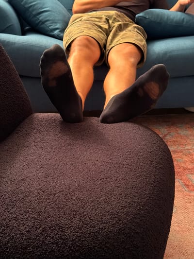 The image shows a person's legs resting on a couch and a large, dark brown, textured object, possibly an ottoman or a large floor cushion. The person is wearing khaki shorts and black socks that have holes in them, revealing parts of their feet. The socks appear worn and stretched. The couch is a deep blue color, with visible seams and cushioning. The background shows a glimpse of a rug with a pinkish-red pattern. The lighting suggests it could be daytime, possibly indoors with natural light. The overall scene appears casual and relaxed, with the person in a reclined position. There is no visible text in the image. The setting appears to be a living room in a home, likely in Glendale, United States of America, given the provided location context.