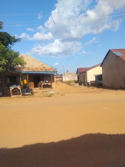 In Mangu Town, Nigeria, this image captures a street scene under a partly cloudy sky. The foreground is dominated by a dirt road. On the left side of the road, a corrugated metal-roofed building with a blue trim appears to be a shop. It has a barred storefront and several wooden tables in front, one displaying a blue tarp-covered item and another with a yellow jerrycan. A motorcycle is parked on the dirt in front of this shop. Further down the street, on the right, is another building with a light-colored facade and a red corrugated metal roof. Adjacent to it is a taller, taupe-colored building with a red corrugated metal roof. A motorcycle is parked in front of this building. In the background, more buildings and structures are visible, along with some distant figures of people walking. Overhead, power lines stretch across the frame.