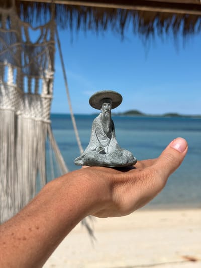 A person's arm and hand hold a small, grey statue of Jesus Christ. The statue is positioned in front of a blurred background of the ocean and beach. The image appears to be taken on a sunny day, with a clear blue sky visible. The presence of a hammock in the foreground suggests a relaxed, vacation-like setting. The shallow depth of field focuses attention on the statue and hand, with the background softly out of focus, creating a sense of peacefulness. There is no visible text in the image.