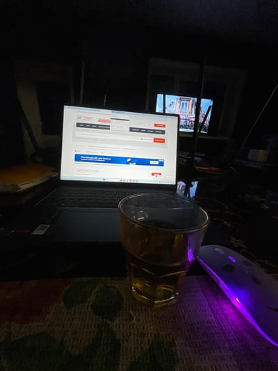 This image depicts a dimly lit indoor scene within the city of Chulym, Russia, observed during evening hours as indicated by the "19:34 МСК" (19:34 Moscow Standard Time) display. The primary subject is a silver-colored laptop, likely a Dell XPS, open and illuminated. Its screen displays a Russian-language website pertaining to an "ЭЛЕКТРОННАЯ ПЛОЩАДКА РОССИИ" (Electronic Platform of Russia), featuring the "PTC" logo and advertisements for services such as "Расчётный счёт для бизнеса" (Current account for business).
In the foreground, an amber-tinted glass tumbler containing a dark liquid is positioned on a patterned, woven surface. To the immediate right of the glass, a wireless computer mouse emits a distinct purple LED light. Further in the background, to the right of the laptop, a second illuminated screen or window displays an image of a building with a balcony, rendered in a bright, colorful style, potentially a European architectural scene. A partial text "ON" is visible on this background display. To the left of the laptop, a stack of papers or books is present on a dark surface. The overall environment suggests a personal workspace during nighttime. No individuals are visible in the scene.