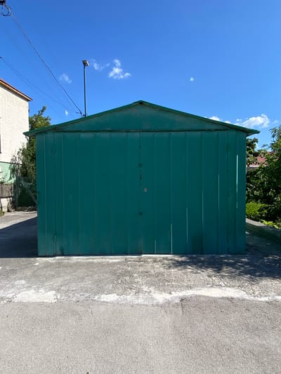 The image depicts a teal-colored corrugated metal garage situated outdoors under a clear blue sky with scattered white clouds. The garage has a peaked roof and double doors. In front of the garage, there is a concrete slab, which is cracked and weathered in places. Beyond the concrete slab, there is an asphalt surface. To the left of the garage, a portion of a light-colored brick building is visible, along with some greenery and a chain-link fence. To the right of the garage, trees and the roof of another building can be seen. A single light pole with a fixture at the top stands behind the garage, to the left of its center. Power lines are visible in the upper left quadrant of the image. The scene appears to be in a residential or semi-industrial area.