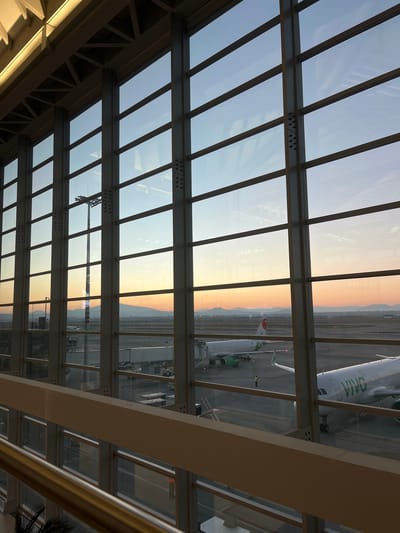 The image depicts an airport terminal interior view looking out onto an airfield during either sunrise or sunset, specifically at Felipe Ángeles International Airport (AIFA) in the vicinity of San Miguel Jaltocan, Mexico.
A large grid-patterned glass facade dominates the scene, supported by dark grey structural steel I-beams and horizontal members. Interior architectural elements, including illuminated ceiling trusswork and a light-colored railing, are visible in the upper and lower portions of the frame.
On the airfield, a tarmac is visible with two white passenger aircraft featuring green livery. The aircraft on the left, bearing "XA-V..." on its tail, is connected to a jet bridge. A small green ground support vehicle is positioned near its forward landing gear. The aircraft on the right, partially visible, clearly displays the word "VIVO" as part of its green fuselage branding.
One individual, wearing a high-visibility vest, is standing on the tarmac between the two aircraft, facing away from the viewer. Various ground support equipment, including white baggage carts and other vehicles, are distributed across the tarmac. A tall floodlight tower stands prominent in the mid-ground.
The sky exhibits a pronounced color gradient, shifting from light blue at the zenith to vivid orange and yellow along the horizon, with distant, hazy mountains silhouetted against the sky. The overall lighting suggests ambient natural light outside, contrasting with internal illumination on the terminal's structural elements.