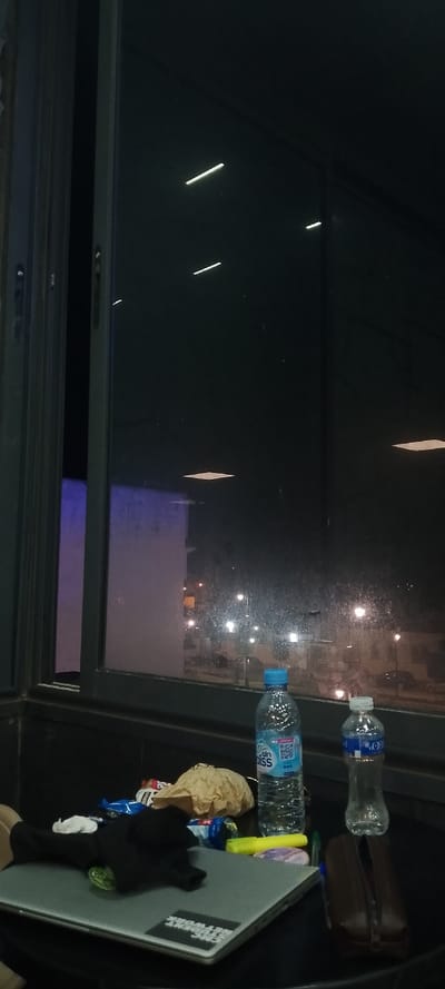 A cluttered desk sits before a large window at night. Two water bottles, snacks in a brown paper bag, and a dark object rest on a laptop. The laptop has a sticker. Outside, streetlights illuminate a cityscape. The window is dirty and reflects interior lights. The room is dimly lit, with a tall, purple-lit wall visible through the window frame.
