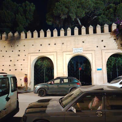 This is a night shot of a street scene in Mechouar Fes Jdid, Morocco. A high, light-colored wall with crenellations runs across the background. The wall has three arched openings with dark, patterned metal gates. A sign is visible above the central arch. Several dark holes are patterned across the wall.
In the foreground are three vehicles. On the left is a white van, partially cut off by the frame. In the center is a dark gray SUV. A black car, with the roof partially visible, is closest to the viewer.
A blurry figure of a person in dark clothing can be seen walking near the white van. Trees are visible behind the wall.