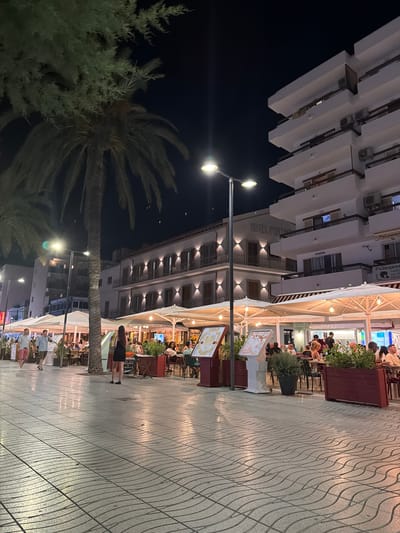 This is a nighttime, outdoor shot of a pedestrian walkway alongside buildings and a restaurant in Sant Antoni de Portmany, Spain. The scene is illuminated by streetlights and the lights from the buildings and restaurant. People are seated at tables outside the restaurant, enjoying a meal or drinks. A few people are walking along the pathway. One woman in a black dress is walking away from the camera. A palm tree is visible in the foreground, and another tree with needle-like foliage is to its left. The buildings appear to be hotels or apartments, with balconies and windows visible. One building has a sign that reads "HOTEL PORTMANY". The ground is paved with a distinctive wavy pattern. The overall atmosphere appears relaxed and social.