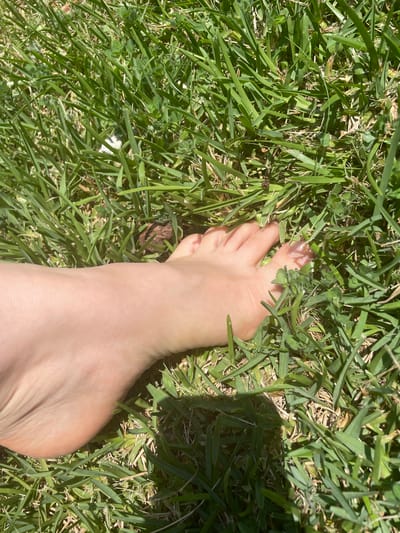 A close-up, high-angle shot shows a bare human foot resting on a patch of green grass. The foot is positioned on the left side of the frame, with the toes pointing towards the right. The skin on the foot is light-colored with visible veins. The grass is dense and appears to be well-maintained, with some patches of dry, brown blades mixed in. Sunlight casts shadows on the grass, creating a dappled effect. There is no discernible location in the city of Albergaria-a-Velha, Portugal, provided within the image itself.