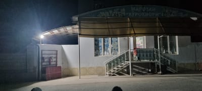 The image shows a storefront at night in what appears to be a village or small town. A brightly lit sign above the entrance reads "МАГАЗИН 'ХУРОКВОРI'" in Cyrillic script. The storefront has large windows and a covered entrance with a yellow awning. There are stairs leading up to the entrance, with a decorative metal railing. To the left of the storefront, there is a white wall with a metal rack holding a red container and a fire extinguisher. The ground in front of the building is paved. The overall scene is illuminated by artificial lights, creating strong contrasts between light and shadow. There are no visible people or vehicles in the immediate vicinity of the storefront. The environment suggests a commercial establishment in a rural or suburban setting. The specific location is identified as Хаваскор, Tajikistan based on the context provided.