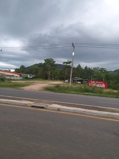 The image depicts a roadside scene in Tambon Pak Phraek, Thailand, under an overcast sky. A multi-lane road dominates the foreground, with a concrete barrier and a yellow line marking its edge. Beyond the road is a grassy area with sparse trees and buildings. On the right, a red sign with Thai script is visible. In the background, a hill or mountain rises above the trees. Power lines stretch across the sky.