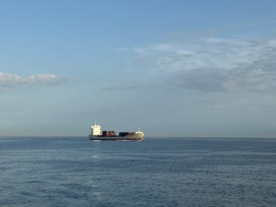 An uncropped, medium-angle, full shot shows a container ship sailing on a vast expanse of calm, blue water under a light blue sky with scattered clouds. The ship, which is white with red accents and loaded with containers of various colors, is positioned in the middle right of the frame. The horizon line is clearly visible and the water has a gentle, rippling texture. The lighting suggests it is either early morning or late afternoon.
