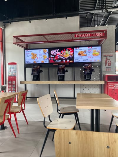 This media file depicts a KFC restaurant interior in Lhoksukon, Indonesia. The scene is dominated by digital menu boards displaying various KFC offerings, including "BURGER COMBO," "SUPER BESAR 2," "SPICY SBESTIE," and "KFC BUCKET," along with their prices in Indonesian Rupiah. There are also smaller screens showing promotional videos or ordering instructions.
The setting includes wooden tables and chairs with red metal frames, characteristic of modern fast-food establishments. The lighting appears to be artificial, suggesting it could be any time of day. The overall atmosphere is clean and functional, typical of a fast-food restaurant focused on efficient service.
Visible text includes the KFC logo, menu item names, prices, and promotional slogans such as "PESAN DISINI" (Order Here), "THE ORIGINAL #BUDAYA BEBERES" (The Original #CleanCulture), and "BERESIN SENDIRI AUTO KEREN" (Clean Up Yourself, Automatically Cool). These slogans promote self-service and cleanliness.
While no people are clearly visible engaging with the ordering system or eating, the presence of chairs and tables implies that customers are expected or have recently been present. The design of the space, with its bright screens and modern furniture, suggests a contemporary dining experience. The overall impression is that of a typical KFC outlet ready to serve customers.