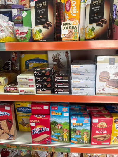 This image captures a detail of product display shelves within a retail establishment, likely a grocery store or supermarket, located in Konstantinovo, Bulgaria. The scene presents three distinct levels of shelving, primarily stocked with packaged food items, predominantly sweets, biscuits, and spreads. The shelves themselves are made of orange-colored metal. On the **top shelf**, from left to right: - A purple-lidded jar of Milka hazelnut spread is partially visible. - Below the Milka jar, a white plastic tub with a red lid, labeled "Eimeti Family Spread," is prominent. - To the right, a yellow carton of "КРЕМ ХАЛВА Слънчогледова" (KREM HALVA Sunflower seed halva) is visible, indicating "gluten-free" and "vegan" characteristics, with a price tag of "1.64€ / 3.20лв". - Dominating the right side are two black boxes of "LAROSHELL WEINBRAND-BOHNE" (Brandy Bean) pralines, described as "Brandy filled chocolates," featuring imagery of grapes and chocolates. A price tag of "2.80€ / 5.48лв" is affixed to one box. The **middle shelf** contains: - On the far left, a box labeled "MY STORY cocoa-coated biscuits" is visible alongside a partially seen box from "Duchars." - In the center-left, a stack of four white boxes labeled "Sweet+ Масленки" (Sweet+ Maslenki - butter biscuits) are arranged, with a partial price tag of "1.00€ / 1.96лв" on the top box. - Next to these, a stack of four black boxes, each labeled "Coffee Cocoa coated biscuits," are present. - Behind these stacked boxes, a large yellow box labeled "Pobeda Черноморец Бонбони" (Pobeda Chernomorets Candies) is partially visible. - On the far right, a tall white box from "TG Biscuits with Tiramisu Flavoured" depicting round chocolate biscuits is displayed, bearing a price tag of "2.91€ / 5.70лв". The **bottom shelf** showcases various wafer products: - On the far left, a red box of "Боровец" (Borovets) wafers is seen, indicating "480g 21 БРОЯ" (480g 21 pieces), priced at "5.50€ / 10.81лв". - To its right, a yellow box labeled "Pobeda Черноморец ОБИКНОВЕНИ ВАФЛИ" (Pobeda Chernomorets Plain Wafers) details "c какао, фъстъци и портокал" (with cocoa, peanuts, and orange), priced at "2.50€ / 4.90лв". - In the center-right, another red box, "ВАФЛА Боровец" (WAFERS Borovets), indicating "375g 15 БРОЯ" (375g 15 pieces), is displayed with a price of "1.80€ / 3.52лв". - Further right, two blue boxes of "Ная ВАФЛИ КЛАСИК" (Naya Classic Wafers) featuring cartoon girl illustrations are visible, one with a price tag of "2.20€ /