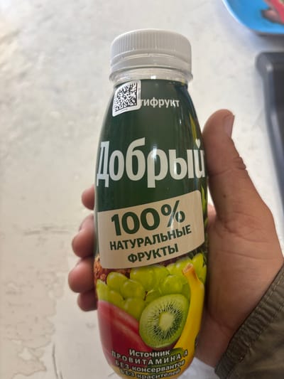 A human hand, appearing to be an adult male's, grasps a green-labeled plastic bottle of "Добрый" (Dobryi) brand fruit juice. The bottle features a white ribbed screw cap and a QR code label with the text "ЦКФРУКТ" near the top. The main label prominently displays "Добрый" in white Cyrillic, followed by "100% НАТУРАЛЬНЫЕ ФРУКТЫ" (100% NATURAL FRUITS). Visuals on the label depict various fruits, including green grapes, a sliced kiwi, a banana, and a red fruit, suggesting a multifruit blend. Additional text states "ИСТОЧНИК ПРОВИТАМИНА А БЕЗ КОНСЕРВАНТОВ БЕЗ КРАСИТЕЛЕЙ" (SOURCE OF PROVITAMIN A WITHOUT PRESERVATIVES WITHOUT DYES). The hand, with a visible light-colored sleeve, holds the bottle from the right side. The background is an out-of-focus, light-toned surface, possibly a table or countertop, with a blurred blue object and a dark, indistinct item in the upper right quadrant. No specific visual cues within the image directly indicate the precise location within the city of Lehvaz, Armenia.