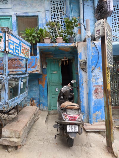 In Jodhpur, India, a scooter is parked in front of a vibrant blue building with peeling paint. To the left of the scooter, a weathered wooden cart with glass panels displays Hindi text, likely an advertisement for a food item called "Dahi Bada." Above the cart, potted plants are arranged on a ledge. A green wooden door is partially open, revealing a dark interior. The ground is composed of concrete and dirt. Various signs and utility boxes are attached to a rusty pole on the right side of the frame. The building's facade features intricate decorative grilles above the windows and door. The overall scene depicts a busy, colorful street in Jodhpur.
