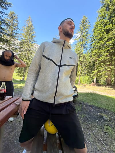 In Govedartsi, Bulgaria, a man wearing a grey Nike hoodie and black shorts sits on a wooden bench outdoors. He is positioned with a yellow ball beneath his legs. Another shirtless man with a black cloth over his head stands to the left of the frame, near a wooden picnic table with a bottle of Coca-Cola on it. Both men are in a wooded area with tall evergreen trees and a bright blue sky. A dark-colored car is visible in the background.