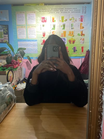The image captures a mirror selfie taken by an individual inside a beverage shop located in Dali City, China. The person, whose upper face is concealed by a light-colored smartphone held in both hands, is visible in reflection. They have long, dark hair, light-colored nail polish (French tips), a ring on their left ring finger, and a dark beaded bracelet on their left wrist, while wearing a dark, long-sleeved garment. The mirror on the right side features a decorative, ornate golden frame. In the background, a large, brightly illuminated poster displays a variety of colorful fruit-based drinks with associated Chinese text, functioning as a menu board. A green potted plant and white decorative shells are situated on a light-brown wooden counter in the midground. A clear plastic-wrapped multipack of bottles is visible on the far left of the counter. The environment is consistent with a commercial establishment specializing in drinks.