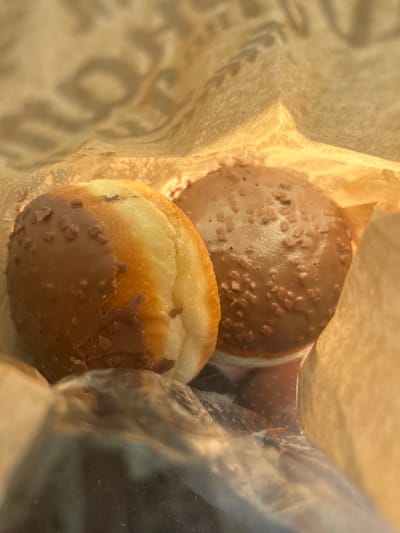 Two donuts are nestled inside a brown paper bag. One donut is completely covered in chocolate frosting and sprinkled with small, crunchy bits, likely nuts or chocolate chips. The other donut is partially dipped in the same chocolate frosting, with the exposed top half showing a golden-brown, bready texture. The donuts are the primary subjects of the image, with the paper bag serving as the setting. The lighting suggests it might be daytime, possibly indoors. There is some text visible on the paper bag, but it is blurry and illegible. The overall impression is one of a simple, perhaps indulgent, treat. The image is taken from a close-up perspective, focusing on the textures and details of the donuts. The crops show the donuts more closely, highlighting the frosting and sprinkles on one, and the bready texture and partial frosting on the other. Another crop shows the interior of the paper bag and the glossy surface of a plastic bag, possibly containing more donuts or another item. The overall scene is static, with no discernible activity or event taking place beyond the presentation of the donuts. There are no people visible in the image. No weather or specific time of day cues are evident. The location context provided is Tsaratsovo, Bulgaria, but this is not visually represented in the image itself.