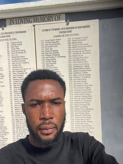 A young man with dark skin, a short beard, and dark curly hair, wearing a dark long-sleeved shirt, is captured in a close-up selfie, looking directly at the camera with a neutral expression. He stands outdoors, partially obscuring a light-colored memorial board. The board's dark text, visible above and around the man, reads "IN LOVING MEMORY OF" at the top, followed by "VICTIMS OF 7TH MARCH, 2010 MASSACRE AT DOGO NAHAWA PLATEAU STATE." Below this, "NAMES OF THE VICTIMS" is listed in multiple columns with corresponding numbers, such as "64. Dagwon Dung" and "127. Dang Daniel Gwom," extending to at least "234. Da Jang Bot Pam." The board is secured by visible fasteners, and a clear blue sky is visible at its top edge. This scene is located in the city of Delimi, Nigeria, commemorating victims from Dogo Nahawa, Plateau State.