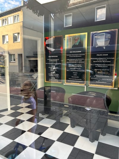 The image shows a storefront window in Waldbröl, Germany, reflecting the exterior of a building and the interior of the store. Inside, promotional displays for "BODYIMPROVER," "DIE VITALIKABINE," and "PELVIC-CHAIR" are visible, detailing various health and wellness treatments. The floor inside the store has a black and white checkered pattern. A young girl wearing a purple shirt and white pants is reflected in the window, looking towards the interior. Two dark-colored, upholstered chairs are also visible in the foreground of the interior. Outside, a building with yellow siding and multiple windows is reflected in the glass. The sky appears blue with some clouds.