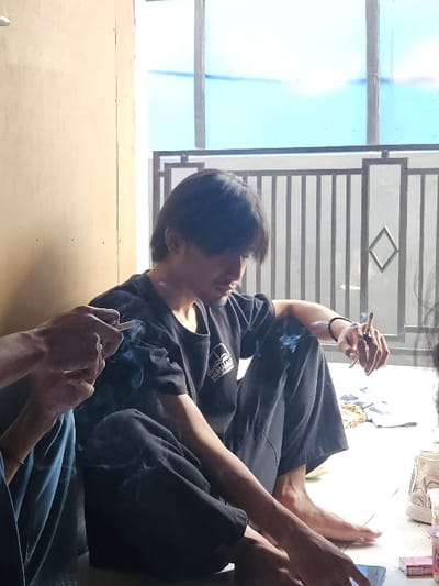 This image depicts three individuals in a semi-enclosed area, likely a residence, in Muka, Indonesia. The central figure is a young male with dark, medium-length hair, wearing a dark t-shirt and dark pants, sitting barefoot on a light-colored tiled floor. He holds a lit cigarette in his right hand, emitting visible smoke, and gazes downwards. Behind him is a dark metal railing with vertical bars and a central diamond design, beyond which a light blue-tinted window structure or sky is visible. To the left, the arm and lower leg, clad in blue pants, of a second individual are visible. This person also holds a lit cigarette, producing smoke, and is positioned near a dark smartphone on the floor. On the far right, dark hair, presumably belonging to a third individual, is partially visible. Adjacent to the third person, various items are scattered on the floor, including crumpled light-colored fabric, a white container, and a small pink rectangular object. A plain light brown wall forms the left background of the scene.