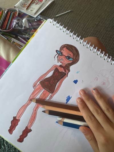 The image depicts an overhead view of an art activity, likely taking place in Valle de Bravo, Mexico, though the specific city location is not visually verifiable within the frame. The primary subject is an open white spiral-bound notebook, containing a colored drawing of a stylized female figure.
The drawing portrays a standing figure with brown hair, tan skin, wearing blue-tinted sunglasses, a dark brown strapless mini-dress with reddish star patterns, a reddish scarf-like accessory on the left shoulder, and reddish-brown boots. To the right of this main drawing, a faint pencil outline of a stylized female head with rosy cheeks and closed eyes is partially visible. Several small, irregular blue marks are scattered on the page near the right side of the main figure.
Five colored pencils (dark brown, light brown, peach/skin tone, white, and dark blue) are stacked diagonally across the lower right portion of the notebook page, partially obscuring the drawn figure's lower legs. A human hand, with light skin tone and extended fingers, rests on the bottom right of the notebook page, covering part of the stacked pencils. A small smudge of blue color is present on the fingernail of the middle finger.
In the upper left background, a pink soft-sided pencil case filled with numerous colored pencils or markers is visible. Behind this, several stacked papers, possibly coloring book pages, are present, one depicting a partially colored cartoon duck-like character. A single white pencil or crayon lies on the background surface. The surface beneath the art supplies appears to be a textured grey material, possibly carpet.