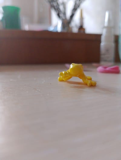 A macro photograph captures a small, yellow, plastic frog toy positioned in a downward-facing, stretched-out posture on a light-colored, textured surface. The frog is the primary subject and is in sharp focus. Behind the frog, a shallow depth of field blurs several objects: a dark brown rectangular box dominates the middle ground, with hints of green and white objects to its left and right. To the right of the box, a blurred pink object is visible. The background is a soft, hazy wash of light colors, suggesting an indoor environment with diffused light. There are no people or animals present in the scene, and no actions or interactions are depicted. The setting does not contain any identifiable landmarks or features that would indicate a specific location within the city of Melitopol, Ukraine, or any other geographical area.