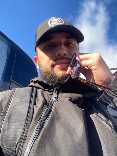 A low-angle, outdoor, waist-up, selfie-style portrait shows a man wearing a black baseball cap with a skull graphic and a black zip-up jacket. He is holding a pair of tortoiseshell sunglasses to his face with his right hand. The man has dark hair, a beard, and is looking at the camera. Sunlight casts shadows across his face and the upper body of his jacket. A dark, reflective surface, possibly a vehicle, is visible to the left of the man. The background consists of a bright blue sky with scattered white clouds. The scene appears to be set in an urban or suburban environment, given the presence of the vehicle. The provided location of Koskolovo, Russia, is a village and is unlikely to be the setting for this image, which seems to be taken in a more developed area.