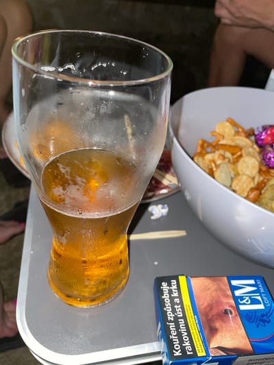 A close-up, outdoor, eye-level shot shows a partially filled glass of beer on a gray table. The beer is a golden amber color with a frothy head and condensation on the glass. To the right of the beer glass is a white bowl filled with a mixture of snacks, including pretzels, crackers, and what appear to be wrapped candies. In the foreground, a pack of L&M Loft Maxi cigarettes is visible, with warning labels in Czech. The background is dark and blurred, suggesting it is nighttime or a dimly lit area. The overall scene suggests a casual gathering or break.