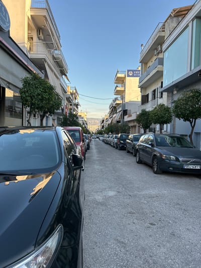This media file captures a static street scene in Koridallos, Greece, depicting a long, narrow residential street lined with parked cars and multi-story buildings on both sides. The image appears to be taken in late afternoon or early morning, indicated by the clear blue sky and the lighting casting distinct shadows. No people are visible.
The street is paved with asphalt, and numerous cars are parked bumper-to-bumper along the right side, extending into the distance. Notable vehicles include a dark grey Volvo SUV with license plate "YZ-4871" prominently visible. Buildings flank both sides of the street, featuring multiple floors, balconies, and awnings. Several air conditioning units are mounted on the building facades. Small trees and bushes are interspersed along the sidewalks, adding greenery to the urban landscape.
Visible text includes:
* A blue sign on one of the buildings (left side, mid-distance) displaying a gold diamond logo and Greek characters, vertically oriented, appearing to read "Π. ΟΥΝ." (P. OUN.).
* A partially obscured sign at the bottom-left edge of the frame, showing what appears to be Greek letters "Π. Υ." (P. Y.), possibly indicating a business like a pharmacy (Φαρμακείο).
