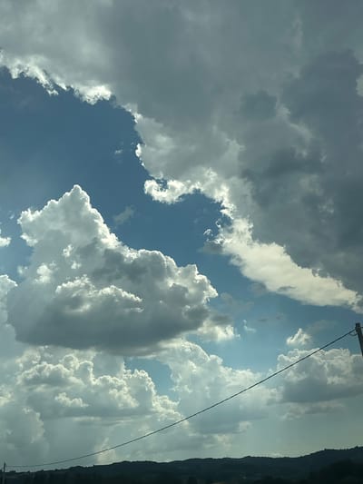 The image captures a dynamic sky over the city of Cannara, Italy. A dramatic display of cumulus clouds, ranging from bright white to dark gray, dominates the scene. The clouds are thick and varied in shape, some appearing fluffy and illuminated by sunlight, while others are dense and shadowed, suggesting an approaching weather system. Patches of clear blue sky are visible between the cloud formations, indicating a transition in the weather. In the lower portion of the image, a dark, silhouetted landscape of rolling hills is visible, with hints of vegetation. A thin, dark power line stretches diagonally across the lower half of the frame, connecting to a utility pole on the far right. The overall impression is one of natural beauty and atmospheric movement, with the sky being the primary subject. There are no people or animals visible in the frame. The scene appears to be captured from an elevated viewpoint, possibly a vehicle in motion or a hillside.