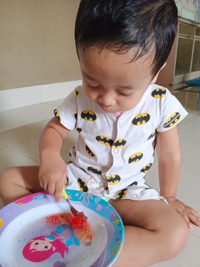 A young child, likely a toddler, is sitting on a tiled floor in what appears to be a residential interior, possibly in Cibungbulang, Indonesia, based on the provided context. The child is wearing a white onesie with a repeating pattern of black and yellow Batman logos. The child is looking down at a colorful plate with a mermaid design, which contains a small amount of liquid and two pieces of red fruit, likely watermelon, on a fork. The child's right hand is holding the fork, and the left hand is resting on their leg. The background shows a neutral-colored wall and baseboard.