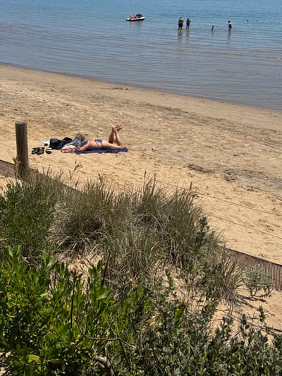 The image captures a sunny day at a sandy beach in Frankston, Australia. The scene is dominated by light-colored sand in the foreground and midground, leading to calm, blue-green water.

In the mid-foreground, a person is seen lying face down on a blue towel on the sand, sunbathing. They are wearing a light-colored bikini or swimwear, with their legs bent at the knees. Nearby, there are some clothes or a bag and a pair of sandals on the sand. A wooden post is visible close to the sunbather.

To the right, a strip of coastal vegetation runs parallel to the shoreline, featuring dry, tall grasses and lush green shrubs. A dark mesh fence or netting separates this vegetation from the open beach.

Further out in the shallow water, several other people are visible. Two individuals stand together, while others are scattered further in the distance, enjoying the water. A small, red and white inflatable boat, possibly a kayak or paddleboard, floats close to the shore on the left side of the frame.

The bright lighting and calm water suggest pleasant weather, likely midday or early afternoon, contributing to a relaxed and leisurely atmosphere. There is no visible text in the image.