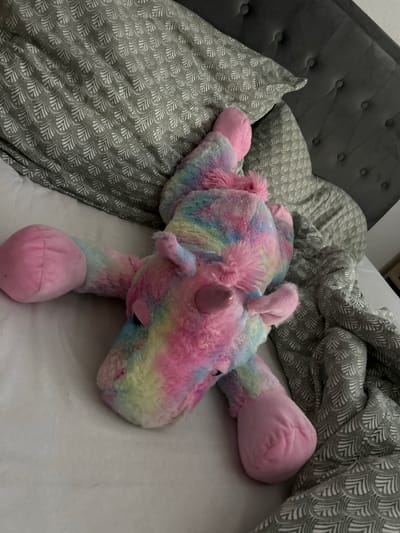 A stuffed unicorn toy with rainbow-colored fur lies on a bed. The unicorn is positioned on its side, with its head facing right and its legs splayed out. The bed has a gray tufted headboard and is covered with pillows and a comforter, both of which feature a repeating gray and white scale-like pattern. The scene is indoors.