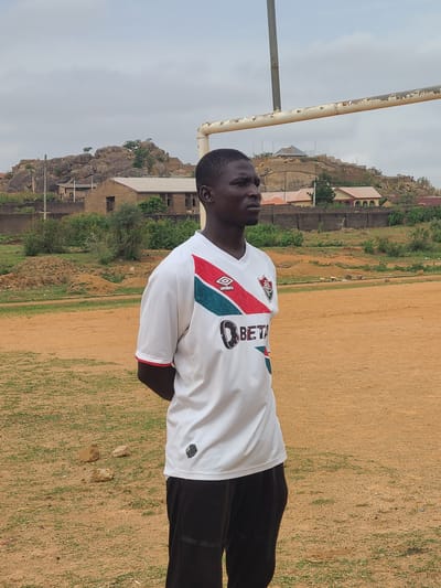 The image shows a person standing on a field. The person is wearing a white jersey with black pants. In the background there are buildings and a rocky hillside. The location is identified as Barikin Jirga Summa, Nigeria.
